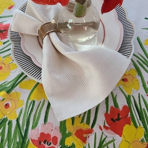 May include: A white linen napkin with a gold napkin ring is placed on a black and white striped plate. A bouquet of red amaryllis flowers in a clear glass vase is on the plate. The plate is on a white tablecloth with a floral pattern of yellow, pink, and red flowers.