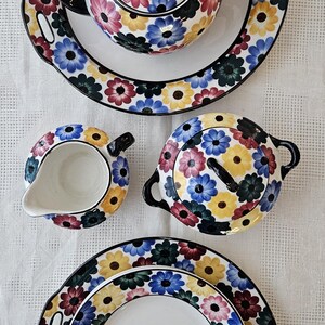 May include: A floral-patterned tea set featuring a teapot, creamer, sugar bowl, and two plates. The set has a white base with a colorful daisy design in yellow, pink, blue, and green. Black handles and rims complete the set.