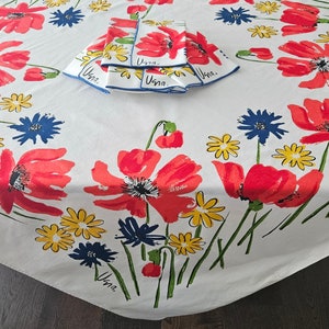 Könnte beinhalten: Weiße Tischdecke mit einem Blumenmuster mit roten Mohnblumen, gelben und blauen Blumen und grünen Stielen. Die Tischdecke hat einen blauen Rand und das Wort "Una" ist in Blau auf der Tischdecke geschrieben.
