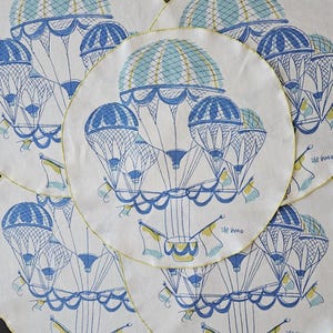 May include: A collection of white circular placemats with a blue and yellow hot air balloon design. The balloons have a detailed basket and flag, with a yellow border. The design is repeated across the placemats.