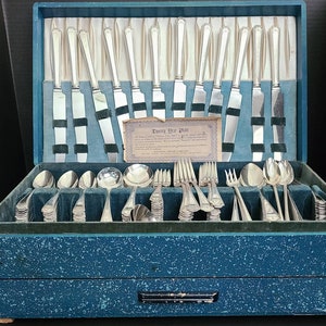 May include: A blue wooden box with a velvet lining contains a set of silver flatware. The box has a label that reads "Twenty-Four Plate" and "International Silver Company".