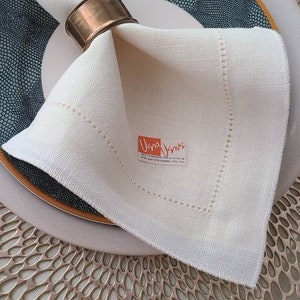 May include: A white linen napkin with a gold napkin ring and a small orange logo that says "Dana Davis" is placed on a blue and gold patterned plate. The plate is on top of a gold metal charger.