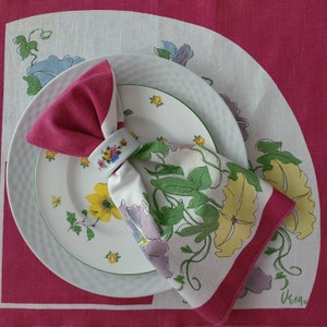 May include: A white napkin with a floral design is folded into a triangle and placed on a white plate with a green rim. The plate is on a pink tablecloth with a floral pattern.
