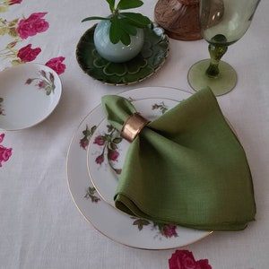 May include: A green linen napkin with a gold napkin ring on a white tablecloth with pink roses. The napkin is folded in a triangle shape and is placed on a white plate with a gold rim.