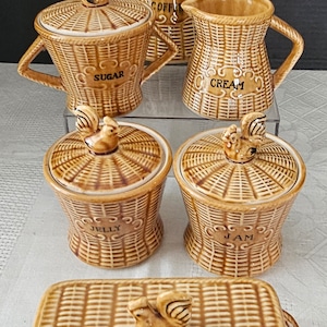 May include: A brown and white ceramic kitchenware set with a wicker pattern. The set includes a coffee pot, creamer, sugar bowl, two small jars, and a butter dish. Each piece has a small rooster figurine on the lid.
