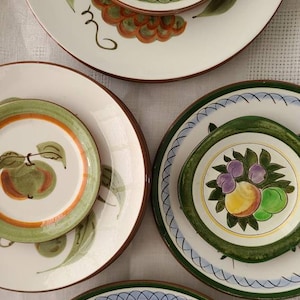 Puede incluir: Cuatro platos de cerámica vintage con fondo blanco y diseños coloridos de frutas y flores. Los platos tienen acentos verdes, azules y naranjas.