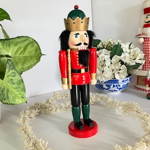 May include: A wooden nutcracker soldier in a red jacket, black pants, and green boots, with a gold crown and a black mustache. The nutcracker is standing on a red base. Other decorative nutcrackers are in the background.