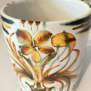 May include: A white ceramic vase with a brown and green floral design. The vase has a wide mouth and a narrow base.