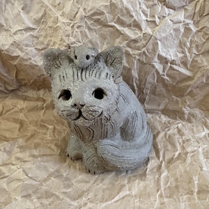 Puede incluir: Una figura de gato de cerámica gris con un pequeño ratón en la cabeza. El gato tiene ojos grandes y abiertos y una superficie texturizada con detalles de purpurina. La figura está sobre un fondo de papel marrón arrugado.