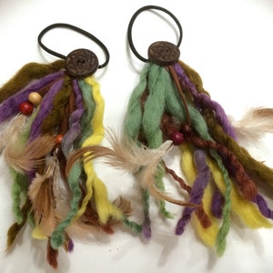 Puede incluir: Dos lazos para el pelo coloridos con botones de madera y largos hilos de lana y plumas en tonos verdes, marrones, morados y amarillos.