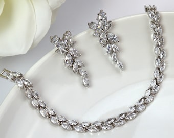 May include: A silver-toned bracelet and matching earrings set. The bracelet and earrings feature a design of marquise-cut clear crystals. The jewelry is displayed on a white surface, with a white flower in the background.