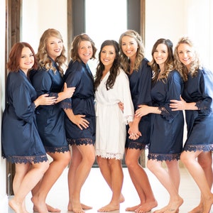 May include: Seven women in navy blue satin robes with lace trim stand in a hallway. The bride is in the center wearing a white satin robe with lace trim.