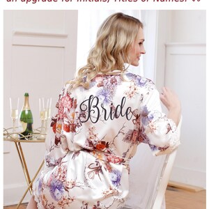 May include: A white satin robe featuring a floral print in shades of red, purple, and brown, with the word Bride printed in black script on the back. The robe has long sleeves with white lace trim at the cuffs and a matching tie belt at the waist.