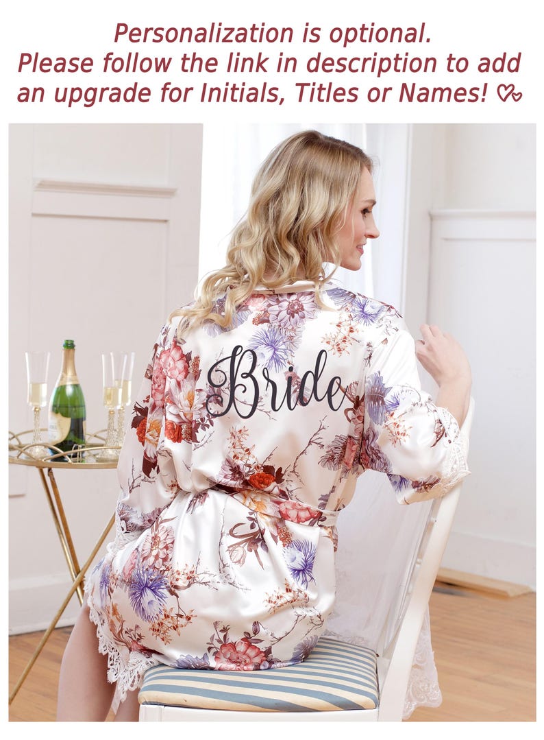 May include: A white satin robe featuring a floral print in shades of red, purple, and brown, with the word Bride printed in black script on the back. The robe has long sleeves with white lace trim at the cuffs and a matching tie belt at the waist.