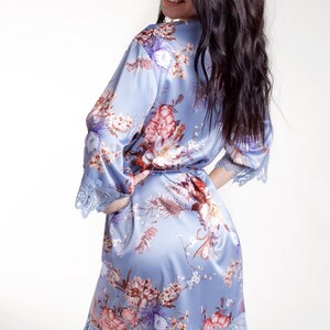 May include: A light blue satin robe featuring a floral print with red, white, and purple flowers. The robe has three-quarter length sleeves and a hemline finished with intricate blue lace trim, along with a matching fabric tie belt at the waist.