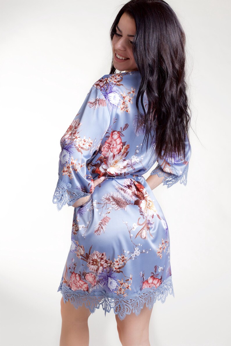 May include: A light blue satin robe featuring a floral print with red, white, and purple flowers. The robe has three-quarter length sleeves and a hemline finished with intricate blue lace trim, along with a matching fabric tie belt at the waist.