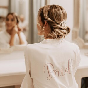 May include: A white bridal robe with the word "Bride" in a soft pink script on the back. The bride's hair is styled in an updo with a decorative hairpiece. The text "Personalization is optional :)" is at the top.