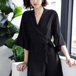 May include: Black wrap-style robe with a V-neckline, tie waist, and ruffled sleeves and hem. The robe is made of a smooth, flowing fabric. The model is standing near a window with greenery in the background.