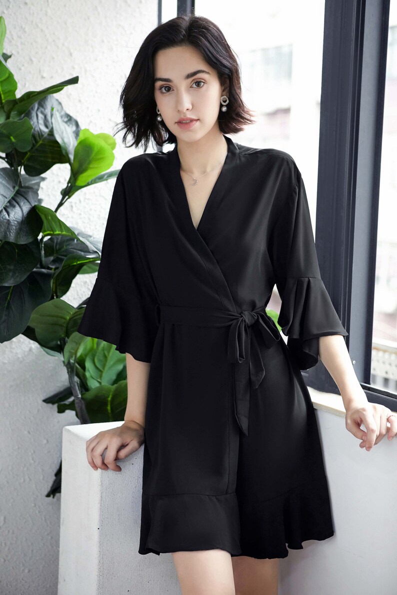 May include: Black wrap-style robe with a V-neckline, tie waist, and ruffled sleeves and hem. The robe is made of a smooth, flowing fabric. The model is standing near a window with greenery in the background.