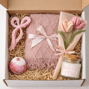 May include: A gift box filled with a pink blanket, a hair clip, a bath bomb, a bouquet of crochet tulips, and a candle. The candle has a label that says "LOVE YOU". The box is lined with shredded paper.