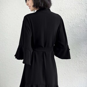May include: A black dress with a matching belt and flared sleeves. The dress has a ruffled hemline and a wrap-style design. The model is wearing the dress, with the back to the camera, against a white wall.