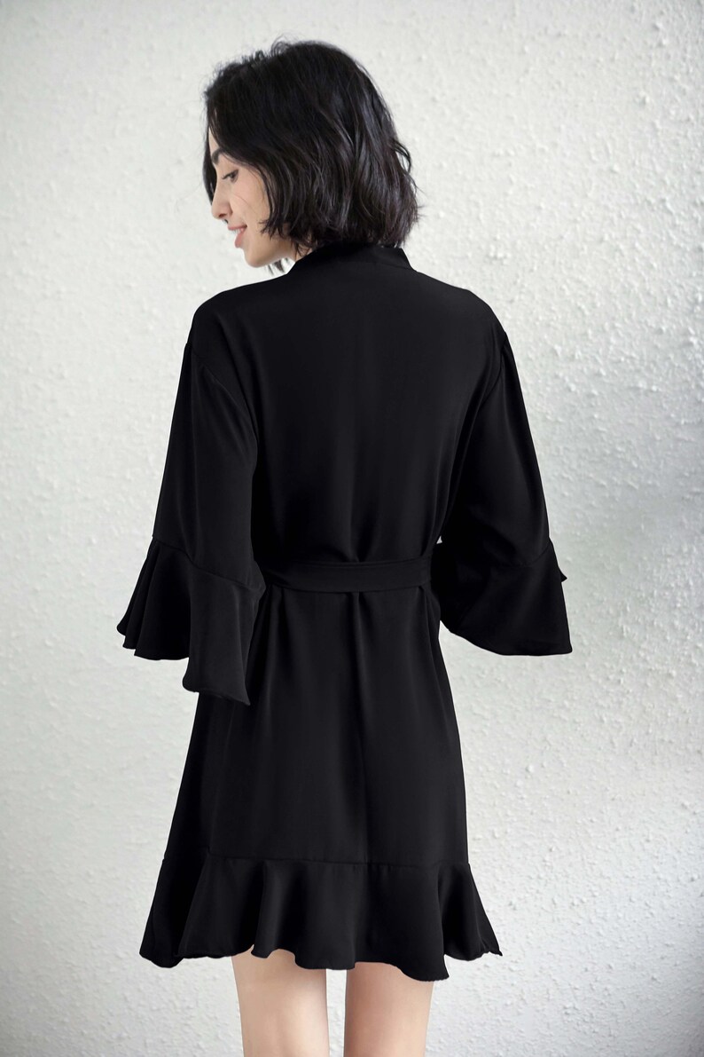 May include: A black dress with a matching belt and flared sleeves. The dress has a ruffled hemline and a wrap-style design. The model is wearing the dress, with the back to the camera, against a white wall.