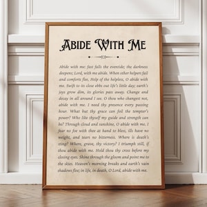 Puede incluir: Una impresión enmarcada con las palabras "ABIDE WITH ME" en una fuente decorativa. El texto del himno está impreso sobre un fondo color crema, enmarcado en un marco de madera marrón. La impresión se muestra contra una pared blanca.