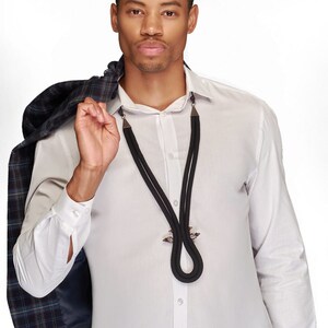 May include: A man wearing a white button-down shirt with a black rope necklace with a decorative metal clasp. He is wearing a plaid jacket over his shoulder.