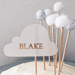 Puede incluir: Pastel blanco con un adorno en forma de nube que dice "BLAKE" en letras doradas. El pastel está decorado con pompones blancos y plateados en palos.