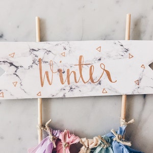May include: A white and gray marble cake topper with the word "winter" written in gold lettering. The topper is on two wooden sticks and has small gold triangle confetti.