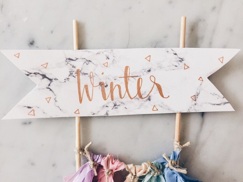 May include: A white and gray marble cake topper with the word "winter" written in gold lettering. The topper is on two wooden sticks and has small gold triangle confetti.