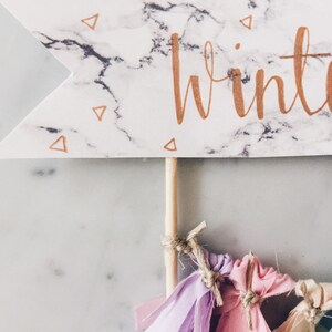 May include: A white marble banner with gold lettering that says "winter". The banner is attached to a wooden stick with colorful fabric tassels hanging from the bottom.
