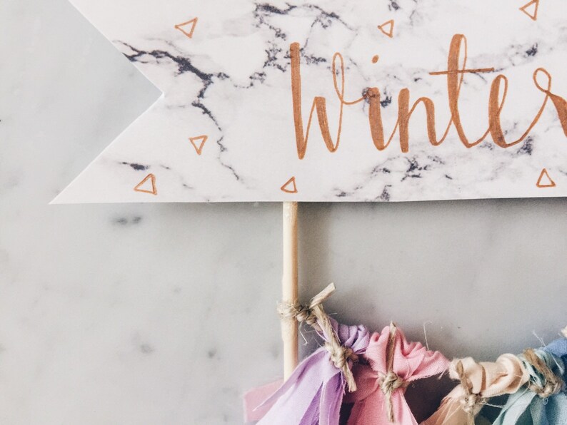 May include: A white marble banner with gold lettering that says "winter". The banner is attached to a wooden stick with colorful fabric tassels hanging from the bottom.
