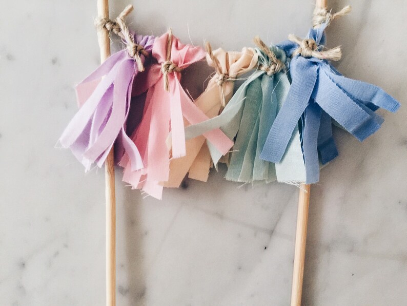 May include: A set of five fabric tassels in shades of purple, pink, cream, blue, and light blue, attached to wooden skewers. The tassels are tied together with twine.