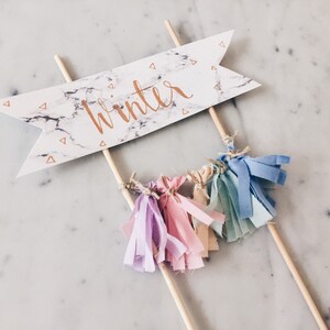 May include: A white cake topper with a marble pattern and the word "winter" written in gold script. The topper has a few colorful fabric tassels hanging from it.