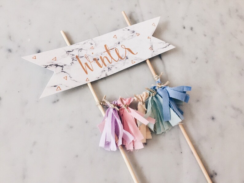 May include: A white cake topper with a marble pattern and the word "winter" written in gold script. The topper has a few colorful fabric tassels hanging from it.