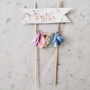 May include: A cake topper with a white marble banner that says "winter" in gold lettering. The banner is attached to two wooden sticks. There are five colorful tassels hanging from the sticks.