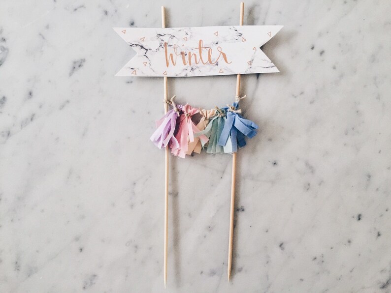 May include: A cake topper with a white marble banner that says "winter" in gold lettering. The banner is attached to two wooden sticks. There are five colorful tassels hanging from the sticks.