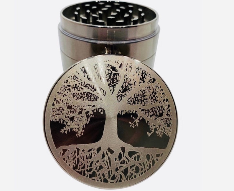 Op de afbeelding: Een zilveren metalen grinder met een levensboomontwerp op de deksel. De grinder heeft vier delen, waaronder een bovendeksel, een maalruimte, een zeef en een onderste kamer.