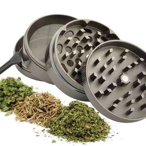 Op de afbeelding: Een zilveren metalen grinder met drie compartimenten. De grinder heeft een zeef en een klein schraapgereedschap. Er liggen drie stapels gedroogde kruiden voor de grinder.