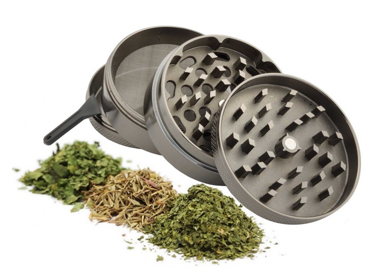Op de afbeelding: Een zilveren metalen grinder met drie compartimenten. De grinder heeft een zeef en een klein schraapgereedschap. Er liggen drie stapels gedroogde kruiden voor de grinder.