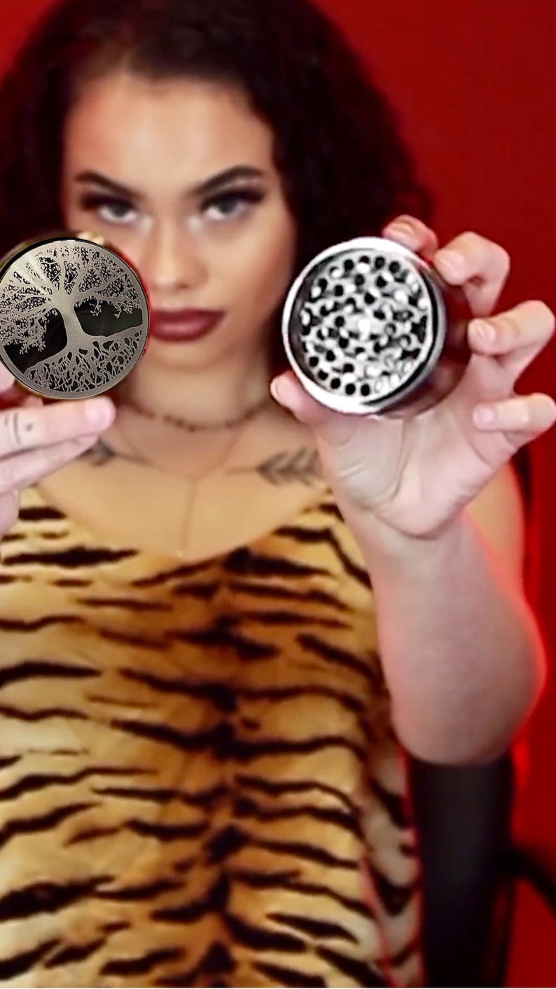 Op de afbeelding: Twee metalen grinders, een met een levensboomontwerp en de andere met een zilveren afwerking en een getextureerd oppervlak.