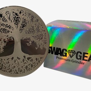 Op de afbeelding: Een zilveren grinder met een levensboomontwerp. De grinder wordt geleverd in een holografische doos met de tekst "SWAG GEAR Premium Grinder".