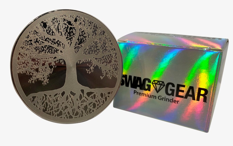 Op de afbeelding: Een zilveren grinder met een levensboomontwerp. De grinder wordt geleverd in een holografische doos met de tekst "SWAG GEAR Premium Grinder".