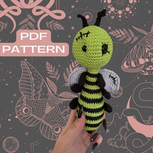 Puede incluir: Un juguete de abeja amigurumi de crochet con una cabeza verde, ojos negros y un cuerpo rayado en negro y verde. La abeja está sosteniendo sus alas hacia los lados. El fondo es gris oscuro con patrones rosas y blancos.