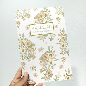 May include: A white planner and journal with a floral pattern in gold and light blue. The cover has the text "RAMADAN PLANNER & JOURNAL" in a gold rectangle.