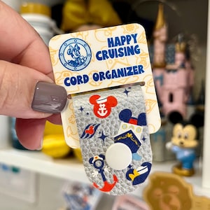 May include: A silver cord organizer with a white snap closure, featuring nautical and Mickey Mouse-themed designs. The card reads "Happy Cruising Cord Organizer" in blue text. The organizer is held in front of a white shelf.
