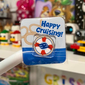 May include: A circular metal pin featuring a red and white life preserver design with a blue Mickey Mouse icon in the center. The pin is mounted on a square backing card with a rope graphic and the blue text Happy Cruising! in a clear plastic sleeve.