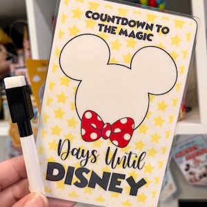 May include: A clear plastic countdown board with the text "Countdown to the Magic" and "Days Until Disney". The board features a white Mickey Mouse head outline with a red polka dot bow. A black and white dry-erase marker is included.
