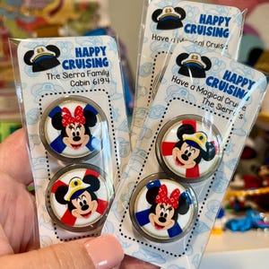 May include: Two clear plastic packages containing circular magnets featuring Mickey and Minnie Mouse characters. The packaging displays the text Happy Cruising, The Sierra Family, Cabin 6194, and Have a Magical Cruise. The magnets have white, red, and blue accents.